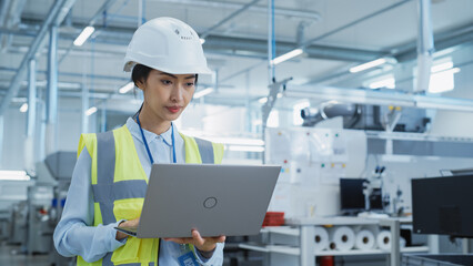 A Focused and Professional Asian Female Engineer in White Hard Hat Standing with Laptop Computer at Electronic Manufacturing Factory. Successful Employee Working on Daily Tasks