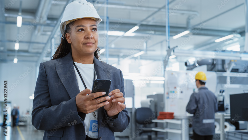 Poster portrait of a black female engineer in hard hat standing and using a smartphone at electronics manuf