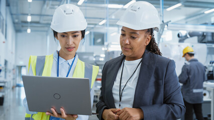 Portrait of Two Female Employees in Hard Hats at Factory Discussing Assignments at Industrial Machine Facility, Using Laptop Computer. Smiling Asian Engineer and African American Technician at Work.