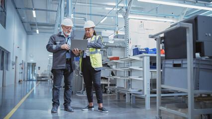 Portrait of Two Heavy Industry Employees in Hard Hats at Factory. Checking and Discussing Industrial Facility, Using Laptop Computer. African American Engineer and Middle Aged Technician at Work.