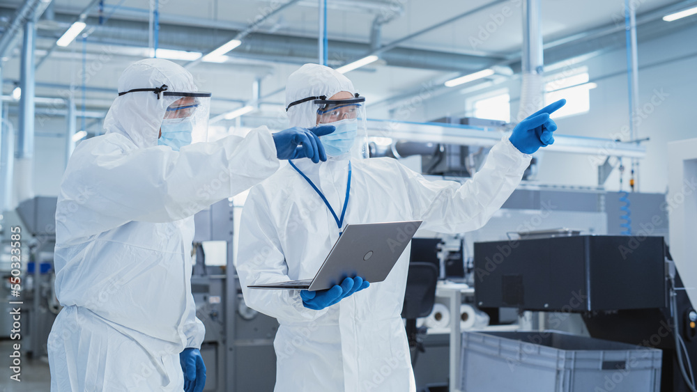 Poster Two Scientists Standing in a Heavy Industry Factory in Sterile Coveralls and Face Masks, Using Laptop Computer. Examining Industrial Machine Settings and Configuring Production Functionality.