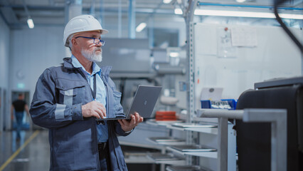 Bearded Middle Aged Chief Engineer Walking in a Factory Facility, Wearing a Work Suit, Stylish Blue Glasses and a White Hard Hat. Heavy Industry Specialist Working on Laptop Computer.