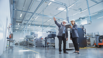 Portrait of Two Heavy Industry Employees in Hard Hats at Factory. Checking and Discussing Industrial Facility, Using Tablet Computer. African American Engineer and Middle Aged Technician at Work.