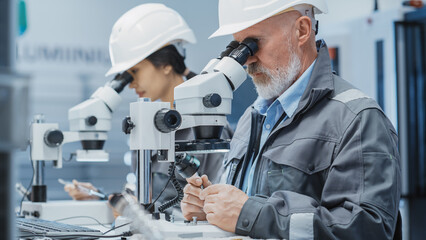 Middle Aged Industrial Scientist and Younger Asian Engineer Working at a Desk in a Factory Facility, Using Microscopes to Work on Small Manufacturing Production Parts and Details.