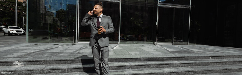 stylish businessman in grey suit holding takeaway drink and talking on cellphone on city street, banner.