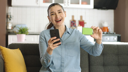 Woman holding smartphone and green box on her sofa, showing a product, smiling and presenting an...