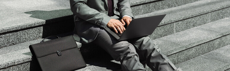 partial view of businessman in grey suit sitting on stairs near black briefcase and typing on laptop, banner.