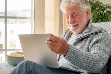 Happy elderly 70s man seated on sofa at home browsing on laptop, older generation and modern tech...