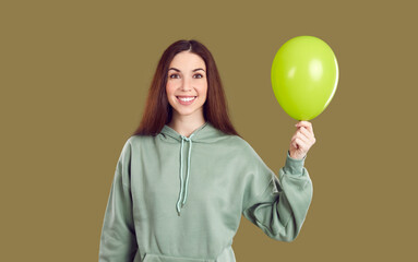 Portrait of joyful young woman holding one inflatable balloon isolated on khaki background. Caucasian young woman in casual hoodie holding light green balloon and smiling while looking at camera.