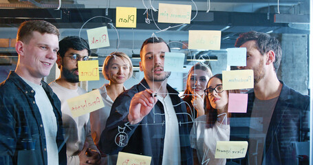 Team of startup project standing in front of board with tasks and to-do notes, drawing chart and...