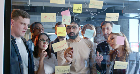 Multiethnic working team making plans for company future and sticking to-do notes. Diverse males and females emplyees brainstorming and planning work for startup project. Management strategy
