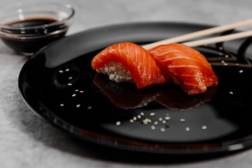 Two servings of sushi with salmon on a black plate and a bowl with soy sauce on a gray background.Close-up
