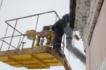 Worker man in overalls in the crane basket remove ice from urban house roof downspout drainage pipe intake at winter day. Roof cleaning, utility service, safety works