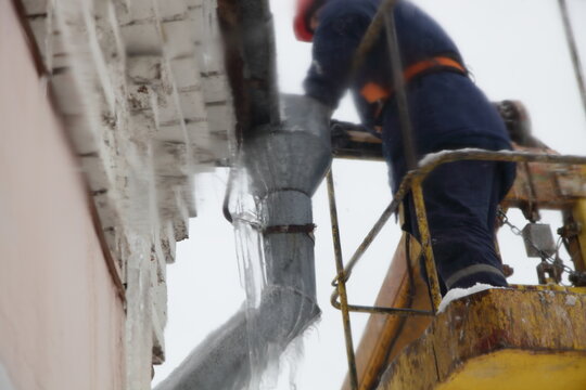 Worker man in overall clothing in the crane basket remove icicles from urban house roof drainpipe intake at winter day. Roof cleaning, utility service, safety works