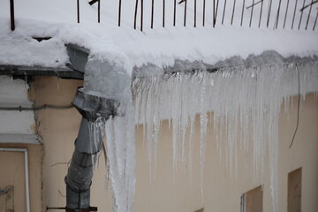 Big icicles on snow covered old roof with downpipe intake on urban house at winter day in Europeю...