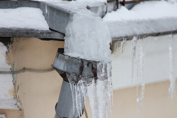 Big icicle on frozen waterspout downpipe intake on house roof at winter day closeup