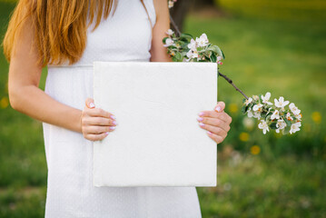 Photo book in a white leather cover in the hands of a woman in spring near a flowering branch of an apple tree - Powered by Adobe