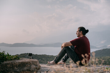 Naklejka na ściany i meble A young man peacefully meets the sunset in a garden with a view of the mountains and the sea.