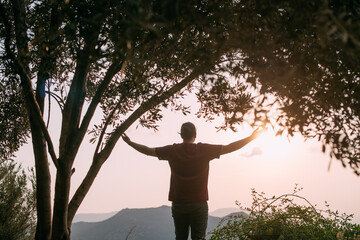 A young man peacefully looks at the sunset, spread his arms, stands on top of a mountain with a view of the mountains and the sea.