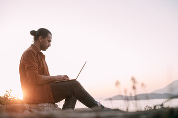 Fototapeta na wymiar A young Caucasian man remotely works with a laptop in a garden on a mountain overlooking the sea and sunset.