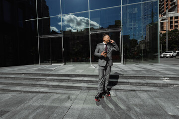 young businessman in roller skates talking on smartphone and holding paper cup near building with glass facade.