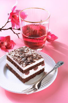 Chocolate cake with sour cherries. Piece of cake on a plate with fork. Sweet dessert on pink background with Spring magnolia blossoms, pink flower decorations. Glass of pink lemonade or rose wine.