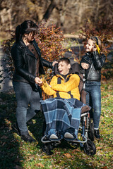 Diversity and inclusion. Happy family, mother, daughter and son teen boy with cerebral palsy spending time together in autumn fall park. Teen boy who uses a wheelchair walking with family - obrazy, fototapety, plakaty