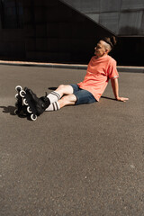 Naklejka premium happy young roller skater with trendy hairstyle sitting on asphalt on city street.