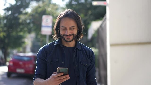 Happy person holding phone walking in city street. Confident hispanic man walks forward looking at cellphone device smiling in movement