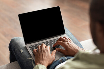 Middle aged african american male sit on sofa watch video, ad and offer, typing on laptop with empty screen