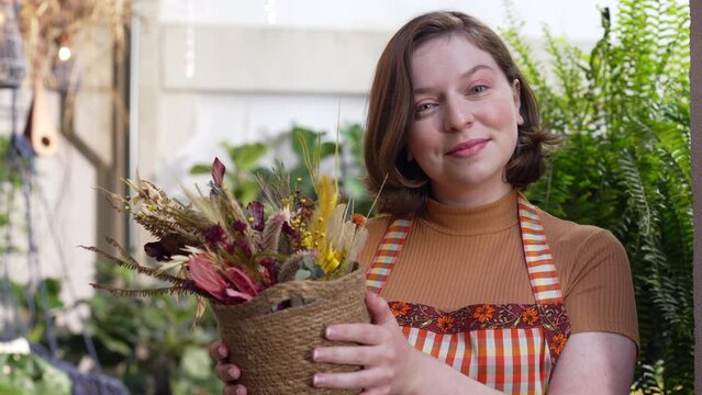 Happy young woman holding bouquet of flower pot showing to camera beautiful arrangement. Portrait girl wearing apron female employee of local small business flower shop