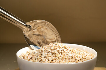 Avena molida siendo vertida en un bol blanco con una cuchara de metal en un fondo aislado. Bol...