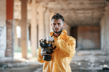 Holding gas mask. Man dressed in chemical protection suit in the ruins of the post apocalyptic...
