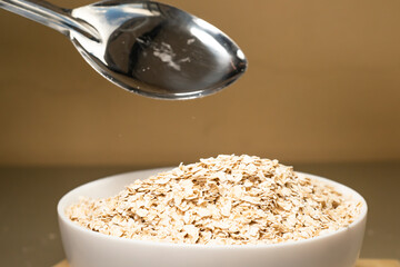 Avena molida siendo vertida en un bol blanco con una cuchara de metal en un fondo aislado. Bol repleto de avena. Plato hondo blanco con avena molida. Plato blanco de cerámica. 