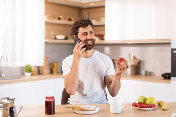 Glad adult caucasian guy with beard in white t-shirt eats sandwich, calls by phone in modern kitchen interior