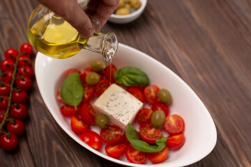 Cherry tomatoes with salted cheese, basil and olives in a deep dish prepared for baking, a man pours olive oil