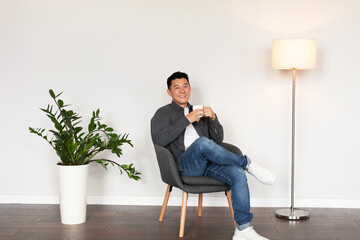 Happy middle aged japanese male enjoy cup of drink, sit in armchair in living room interior with...