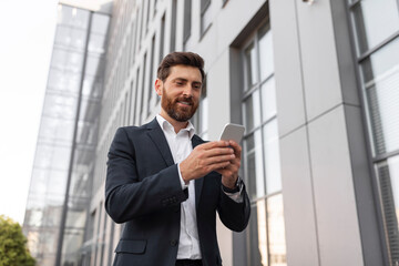 Happy confident young european male boss with beard in suit typing, chatting on phone near building