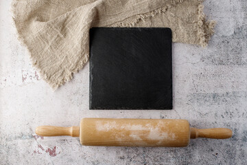 Empty black slate board with linen napkin and wooden rolling pin on stone table. Recipe or menu...
