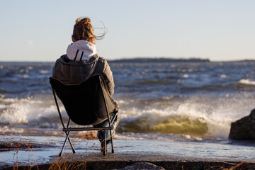 A woman sits on a folding camping chair and looks at the seascape.