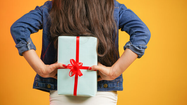Closeup of a girl hand hiding a gift box behind her back, handmade present wrapped in paper, woman with gifts isolated over color background
