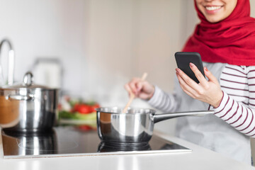 Cropped of woman in hijab cooking and using smartphone