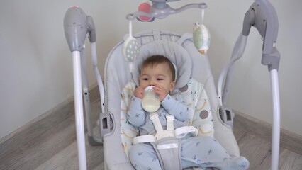 A baby drinks milk from a bottle while swinging in a cradle swing. An automatic baby swing swings a little boy. Closeup.