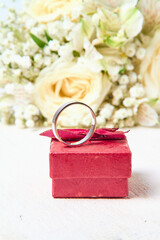 A silver ring stands on a red gift box and a bouquet of flowers on a white textured tree background, holiday