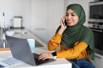 Telecommuting. Female In Hijab Using Laptop And Talking On Cellphone At Home