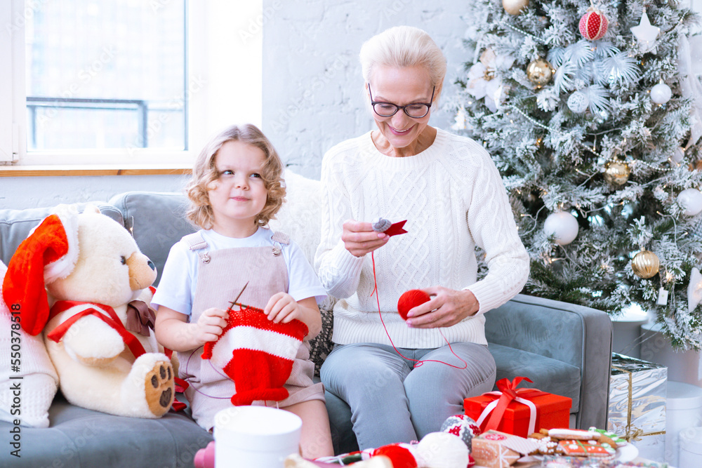 Wall mural Christmas family grandmother and granddaughter sit on sofa near Christmas tree and preparing handmade New Year gifts, pretty girl and active happy senior woman knitting and sewing toys in apartments