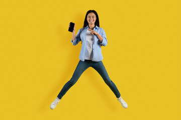 Fototapeta na wymiar Satisfied cute millennial asian female student jump, freeze in air, point finger at smartphone with blank screen