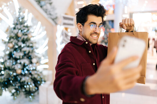Handsome bearded young man looking at camera taking selfie on phone posing with paper bags with gifts standing in hall of celebrate shopping mall, with beautiful xmas decorations in Christmas eve.