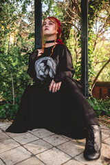Naklejka premium Young red haired girl with red hair scarf and dark clothes and skirt in a old centuries gazebo in the park in the afternoon