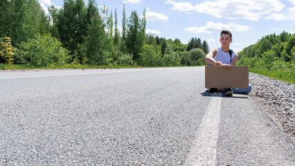 A young man is hitchhiking with a cardboard sign in his hands. Space for text. Passion for travel, the concept of auto travel. A teenager tries to stop a car on the road with an empty cardboard sign